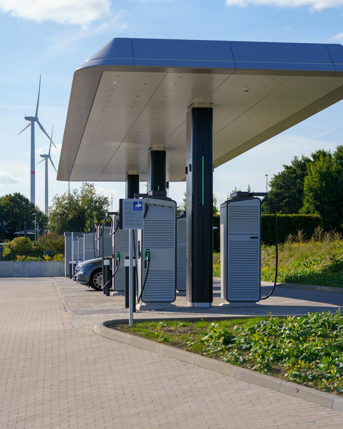 Elektroauto-Ladestationen unter einem Vordach mit Windrädern im Hintergrund.