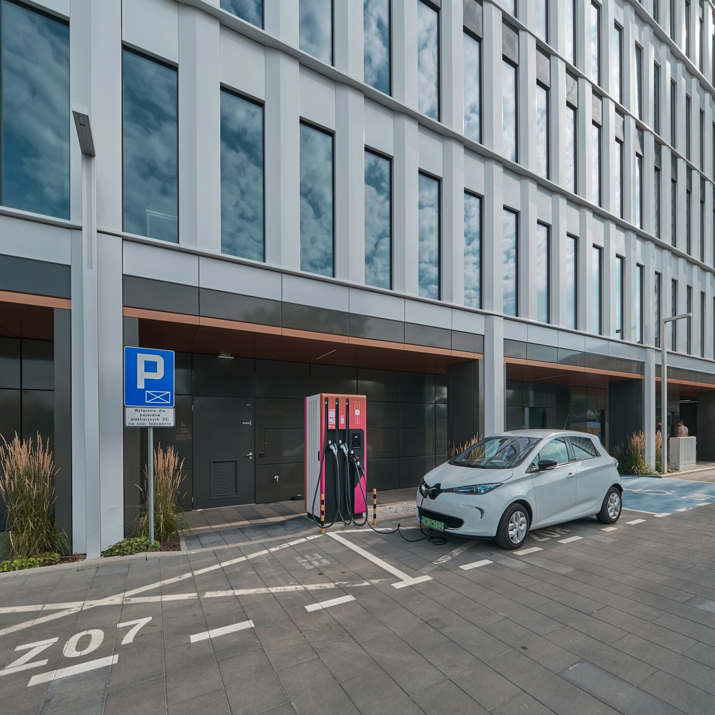 Graues Elektroauto lädt an einer Ladestation in der Nähe eines Bürogebäudes.